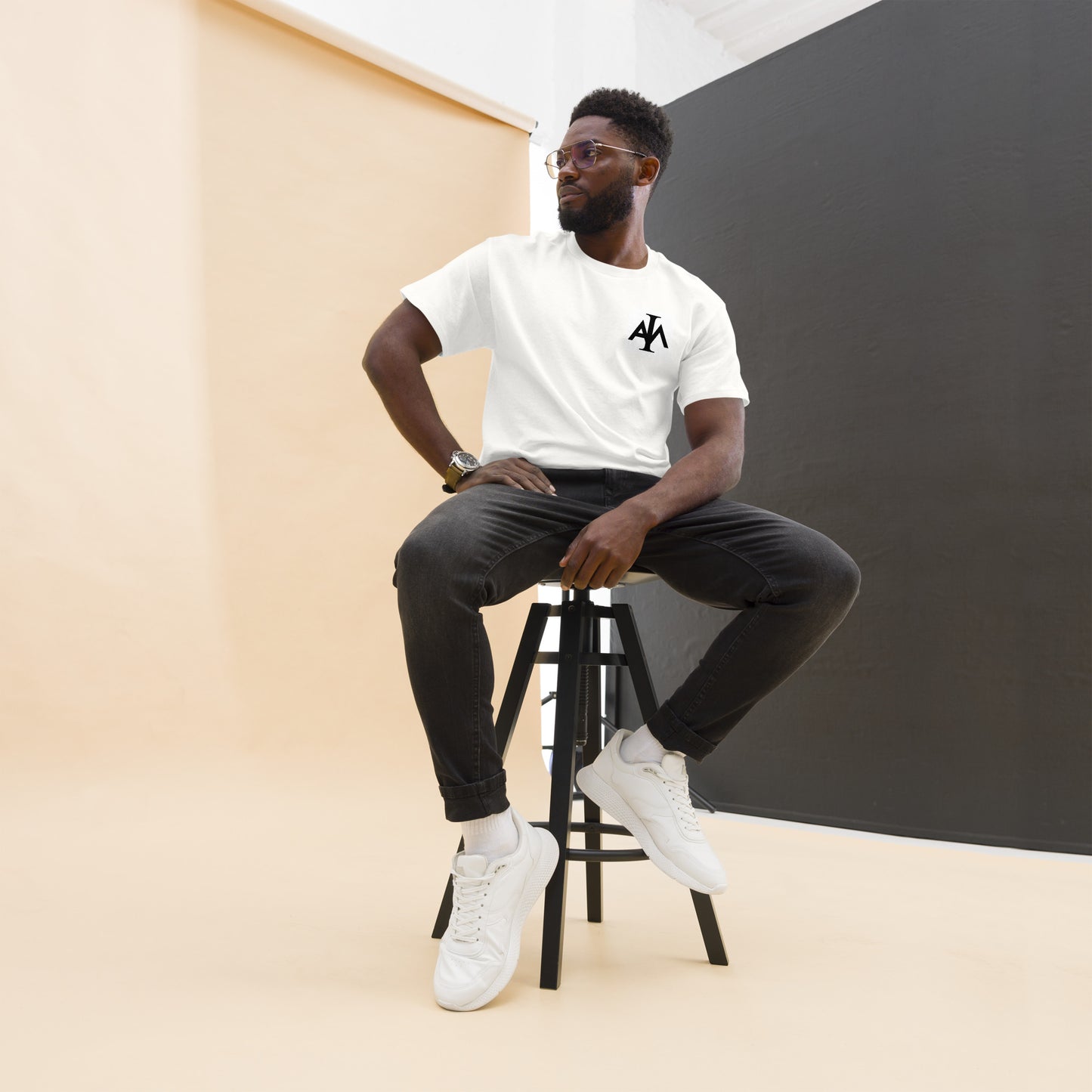 Man sitting on a stool wearing a white t-shirt with a logo, black pants, and white sneakers against a plain background.